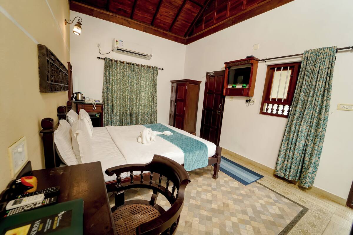 A hotel room with a bed, a desk with a chair, a wardrobe, an air conditioning unit, and patterned curtains.