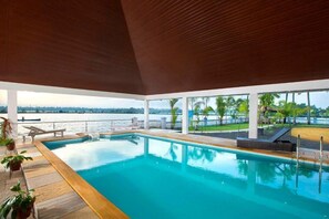 An indoor swimming pool with a view of the water body outside, surrounded by potted plants and lounge chairs. - Indriya Sands (Kochi)