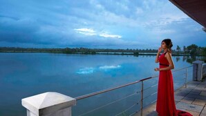 Une femme vêtue d’une robe rouge se tient près d’un lac paisible, bordé d’une rambarde, sous un ciel nuageux.