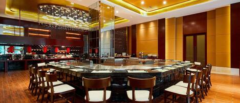 A spacious dining area with a large round table surrounded by chairs, a modern chandelier, and a bar area with red accents.