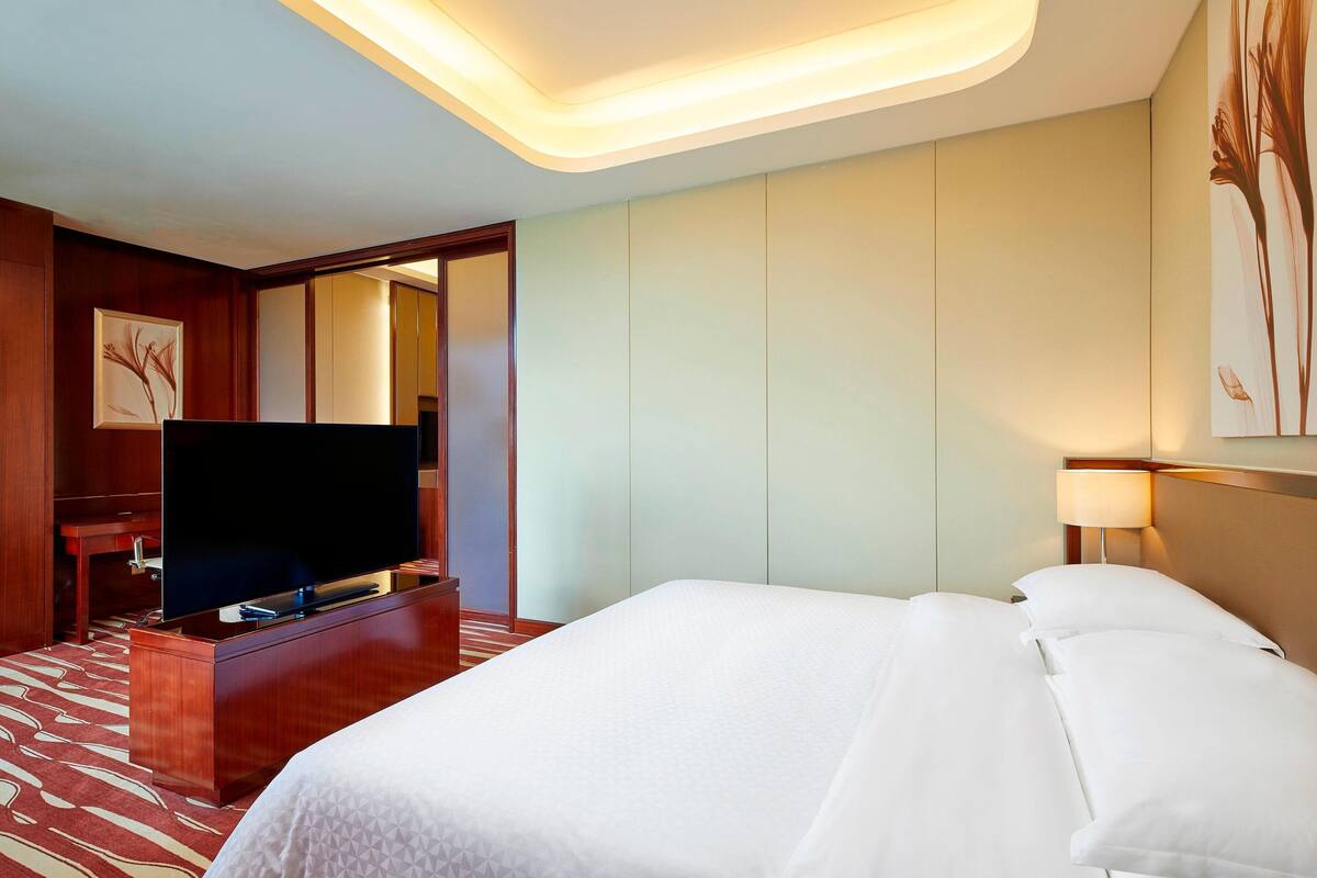 Ein Hotelzimmer mit einem großen Bett, einem Flachbildfernseher auf einer Holzkommode, einer Nachttischlampe und einem an der Wand hängenden Kunstwerk.