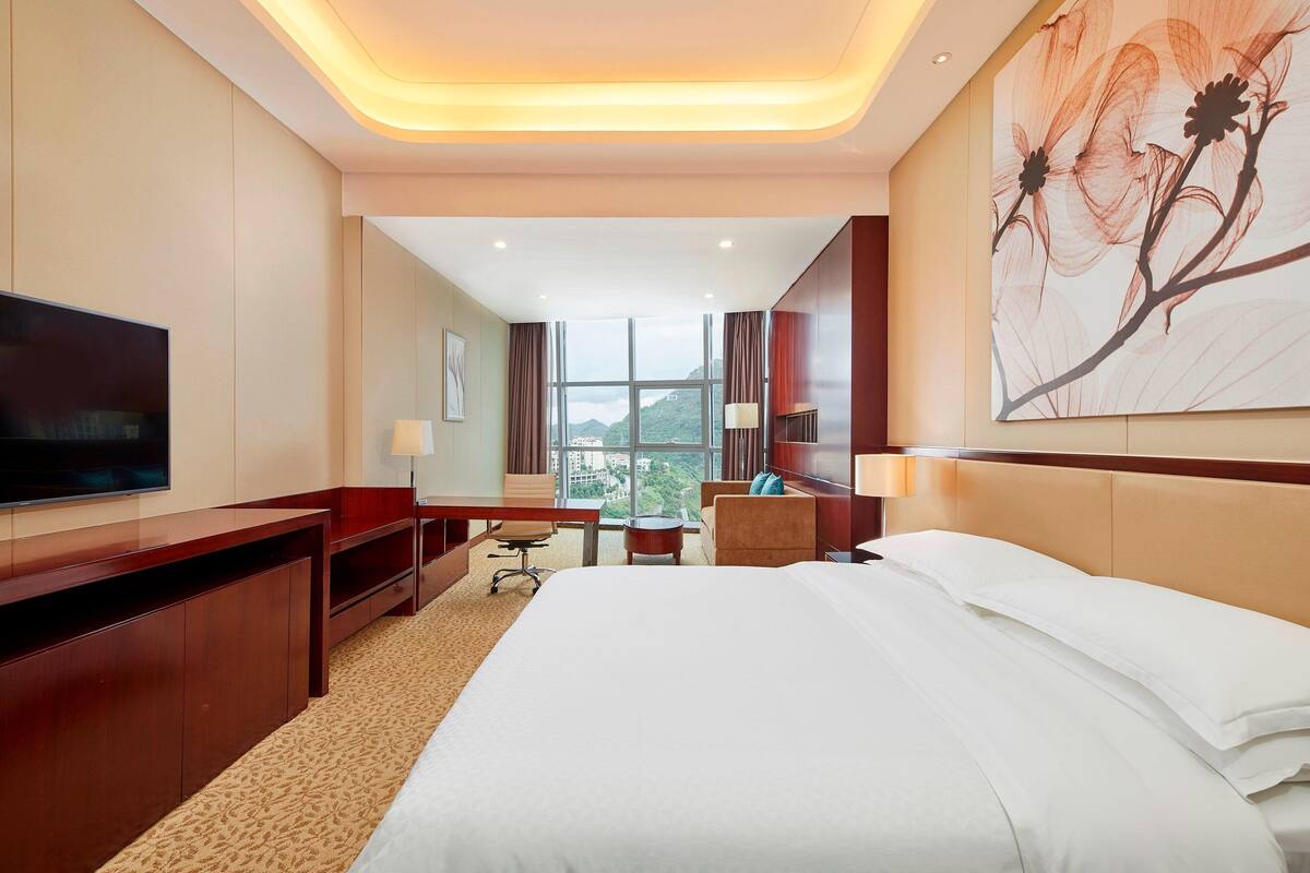 Ein Hotelzimmer mit einem großen Bett, einem Schreibtisch, einem Sessel, einem Fernseher und einem Fenster mit Blick ins Grüne.
