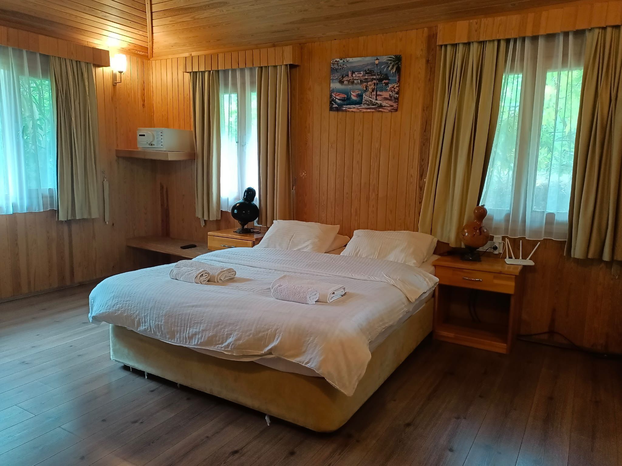 A wooden-paneled room with a large bed, a nightstand, a wall-mounted lamp, and a painting.