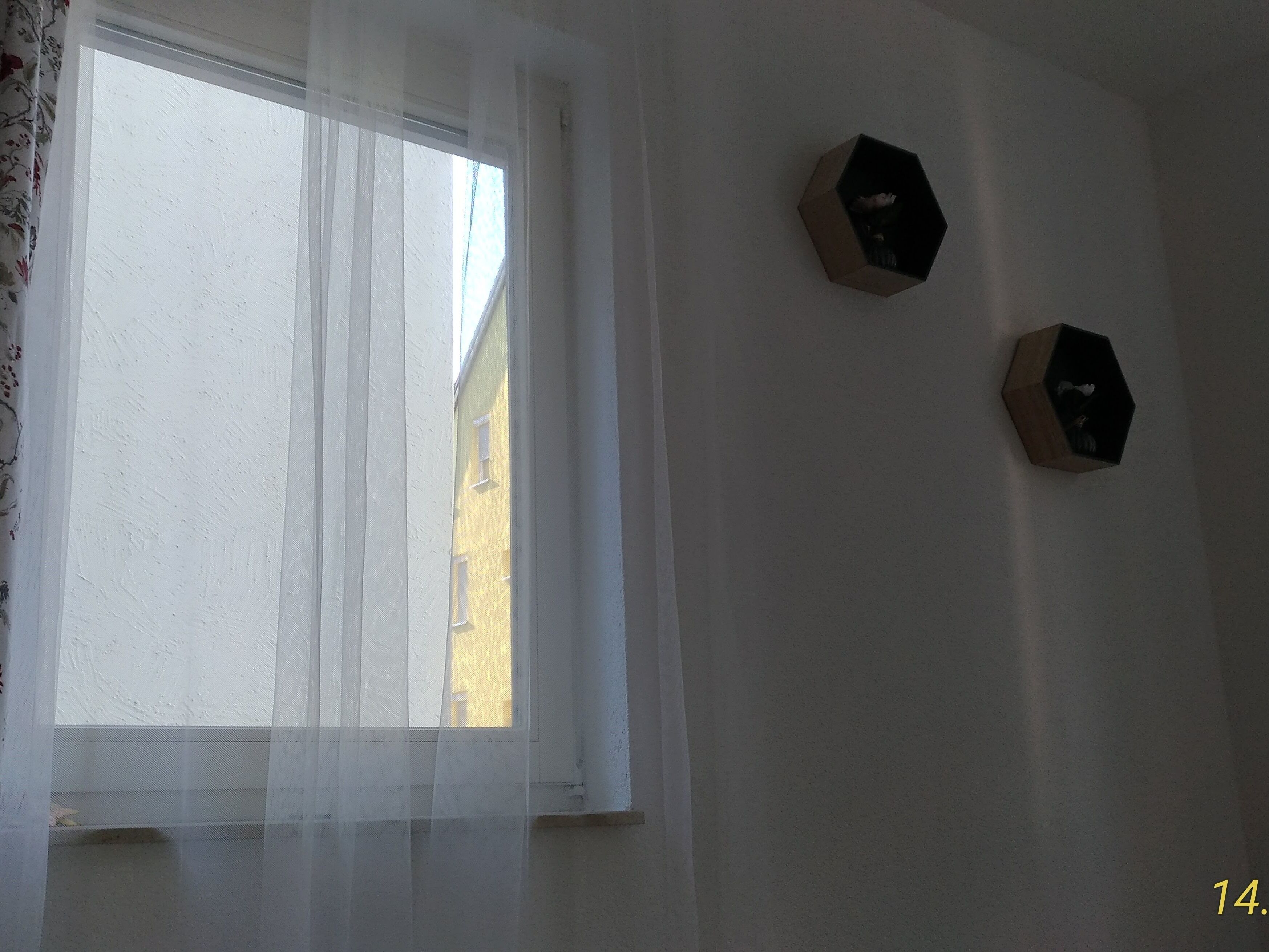 Quarto com janela coberta por cortinas translúcidas, duas arandelas hexagonais e um edifício visível lá fora.
