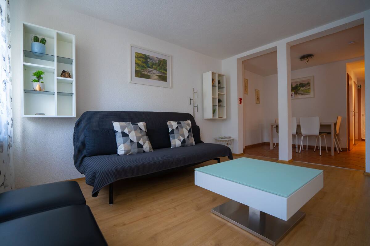 En moderne stue med en mørk sofa, et sofabord i glas og en hvid reol med planter.