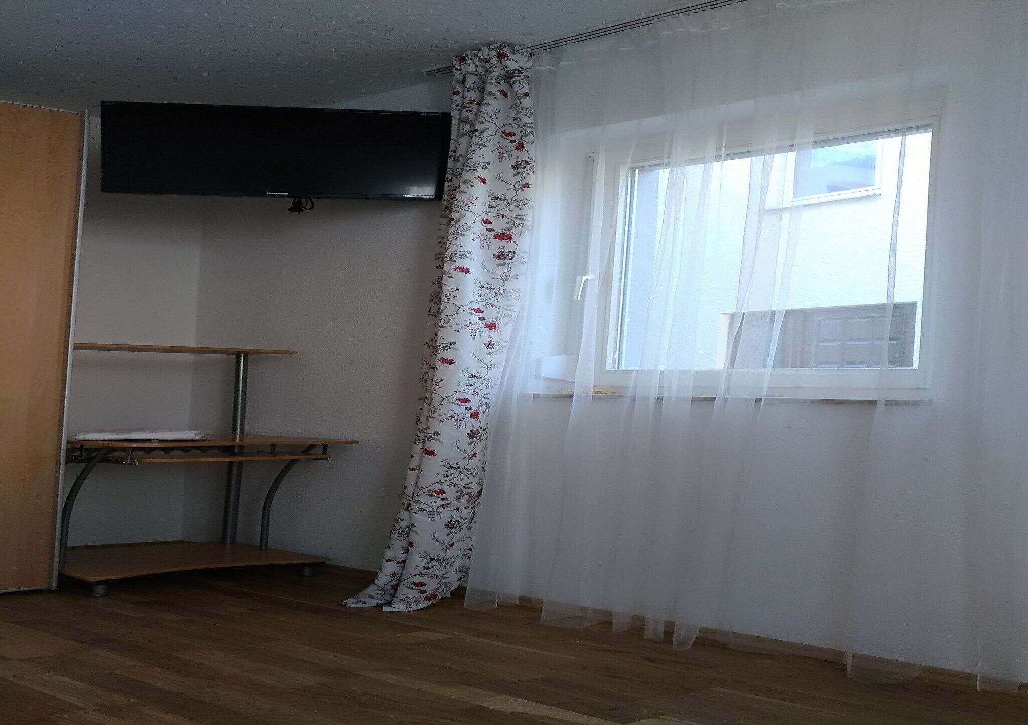 A room with a wooden floor, a window with sheer curtains, a mounted flat-screen TV, and a small table with a shelf.