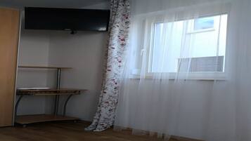 A room with a wooden floor, a window with sheer curtains, a mounted flat-screen TV, and a small table with a shelf.