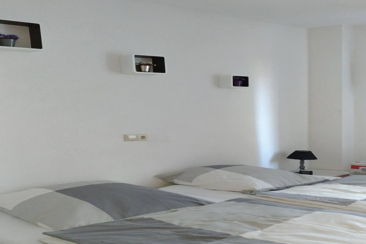 Et soveværelse med to senge, et natbord med lampe, en lille hylde med en potteplante og en fastgjort lampe på væggen.