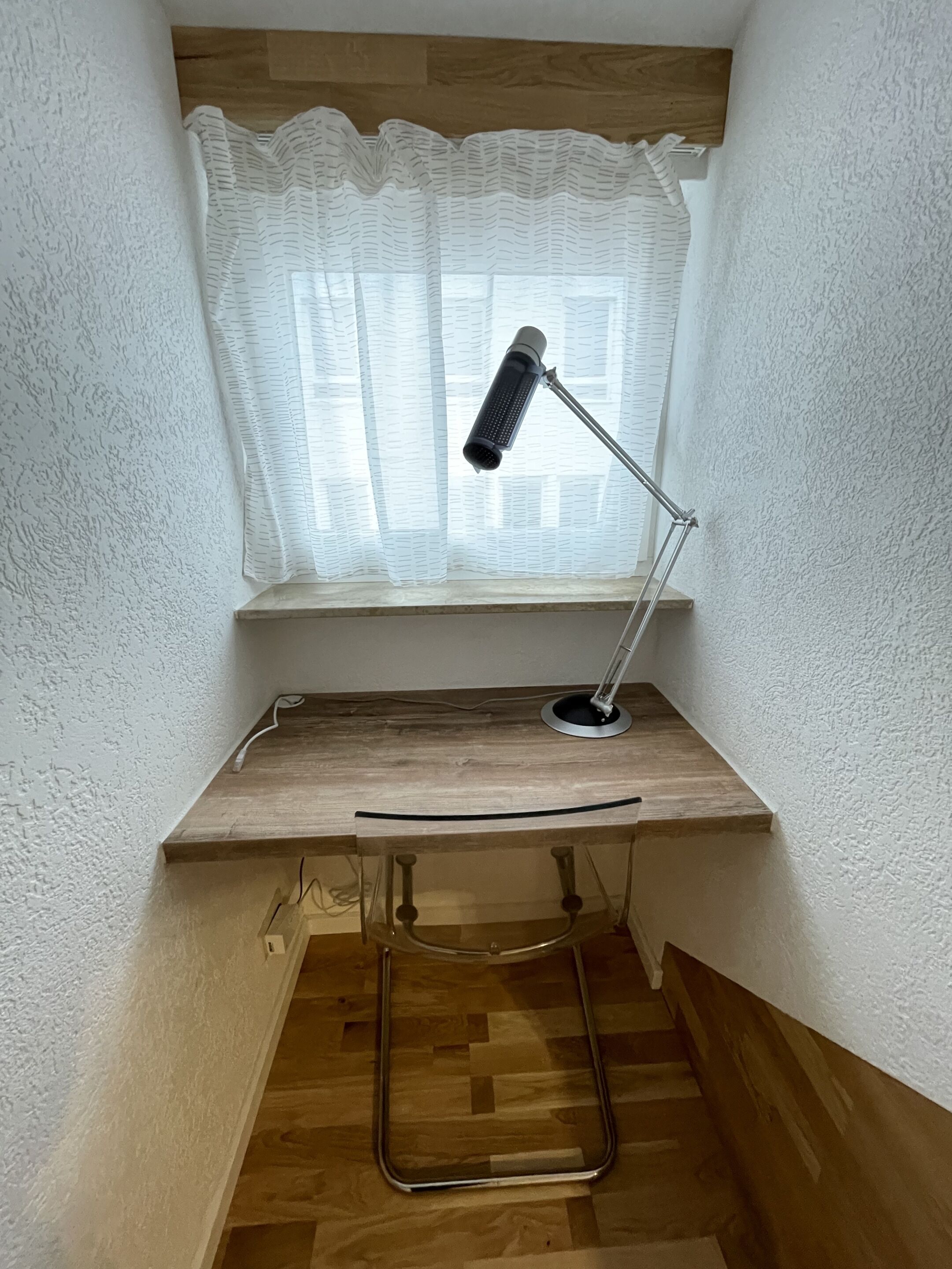 Een bureau in de hoek, een lamp, een raam met een witte gordijn en een houten vloer.