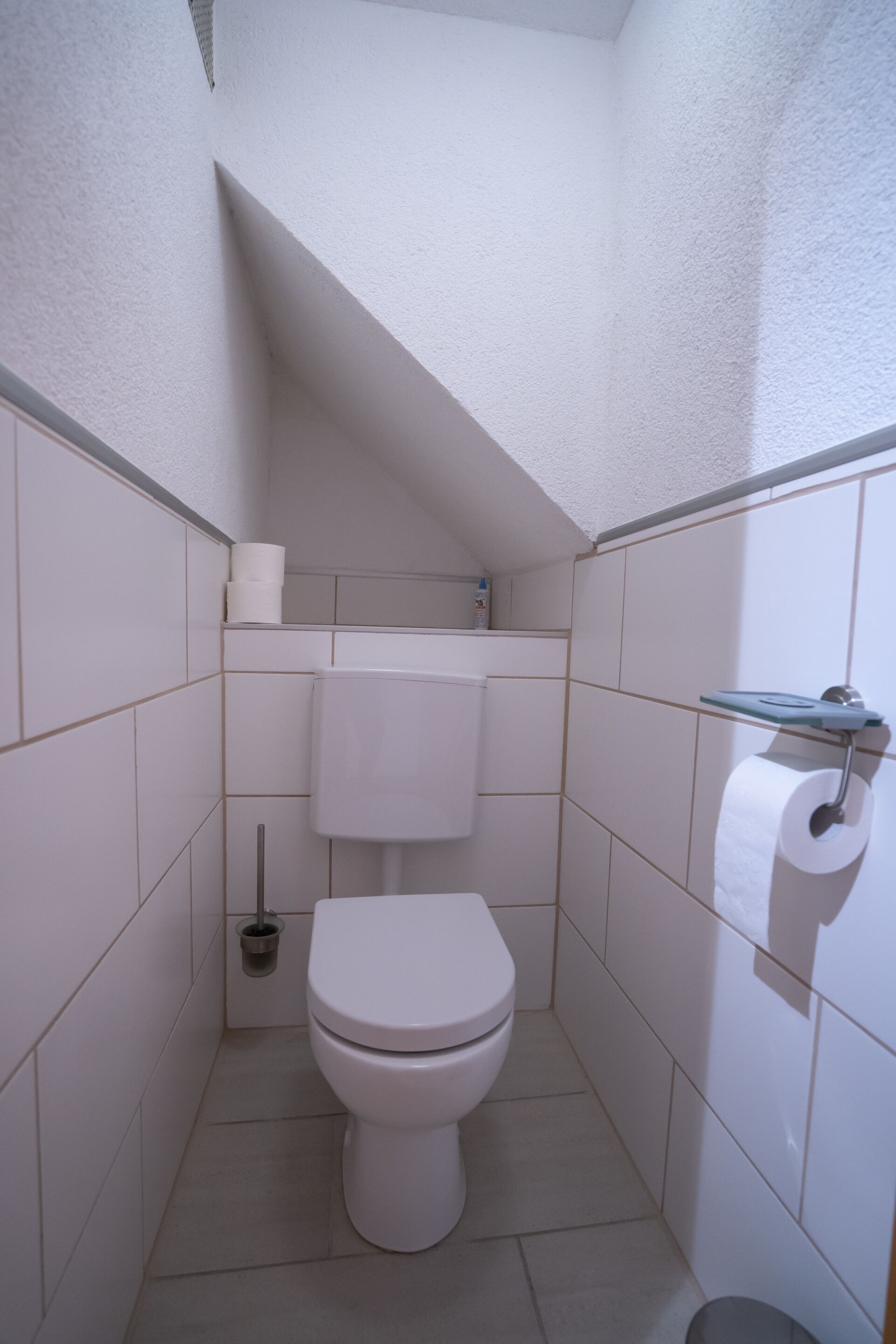 A small, white-tiled bathroom with a toilet, a toilet paper holder, and a hand dryer.