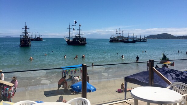 Vista de la playa con varios barcos grandes amarrados en el agua, gente nadando y tomando el sol, y un pequeño kiosco que vende refrescos.