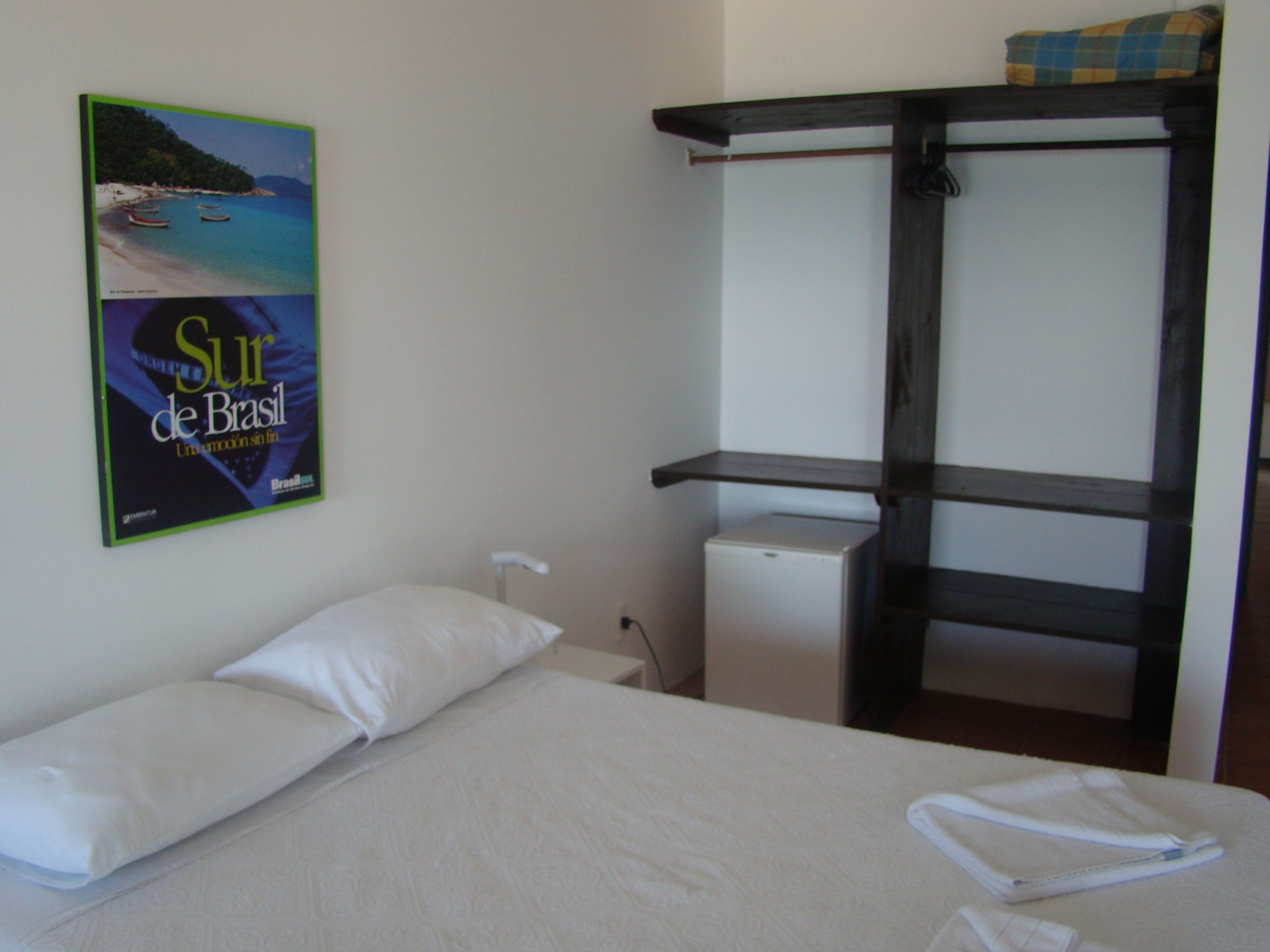 A hotel room with a bed, a small white refrigerator, and a shelf with a towel.