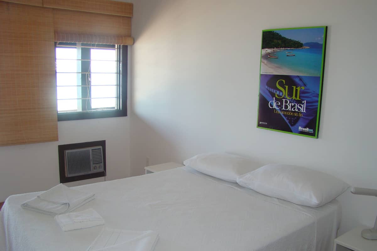 A neatly made bed with white linens and pillows, a window with blinds, a wall-mounted air vent, and a framed picture of a beach scene.