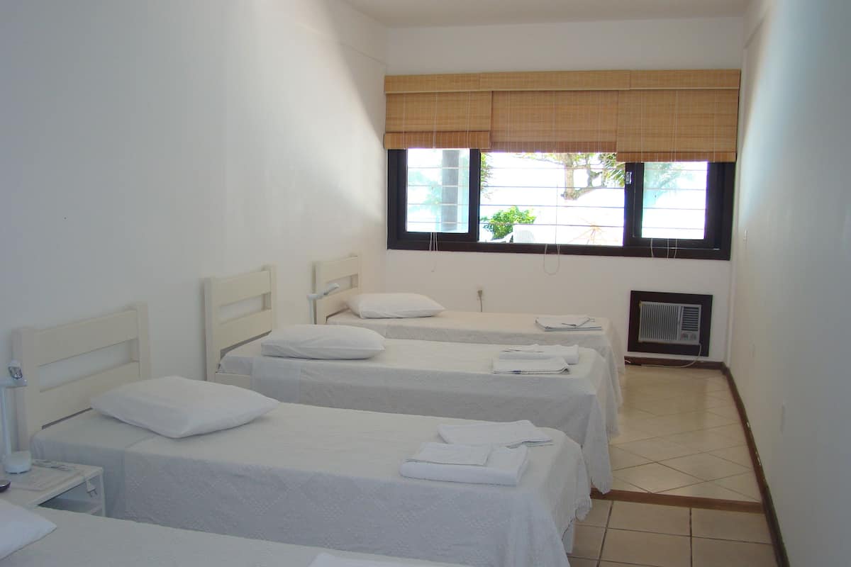 A hotel room with three single beds, each with a pillow and a folded towel. There is a window with a blind, a radiator, and a small table with a lamp.