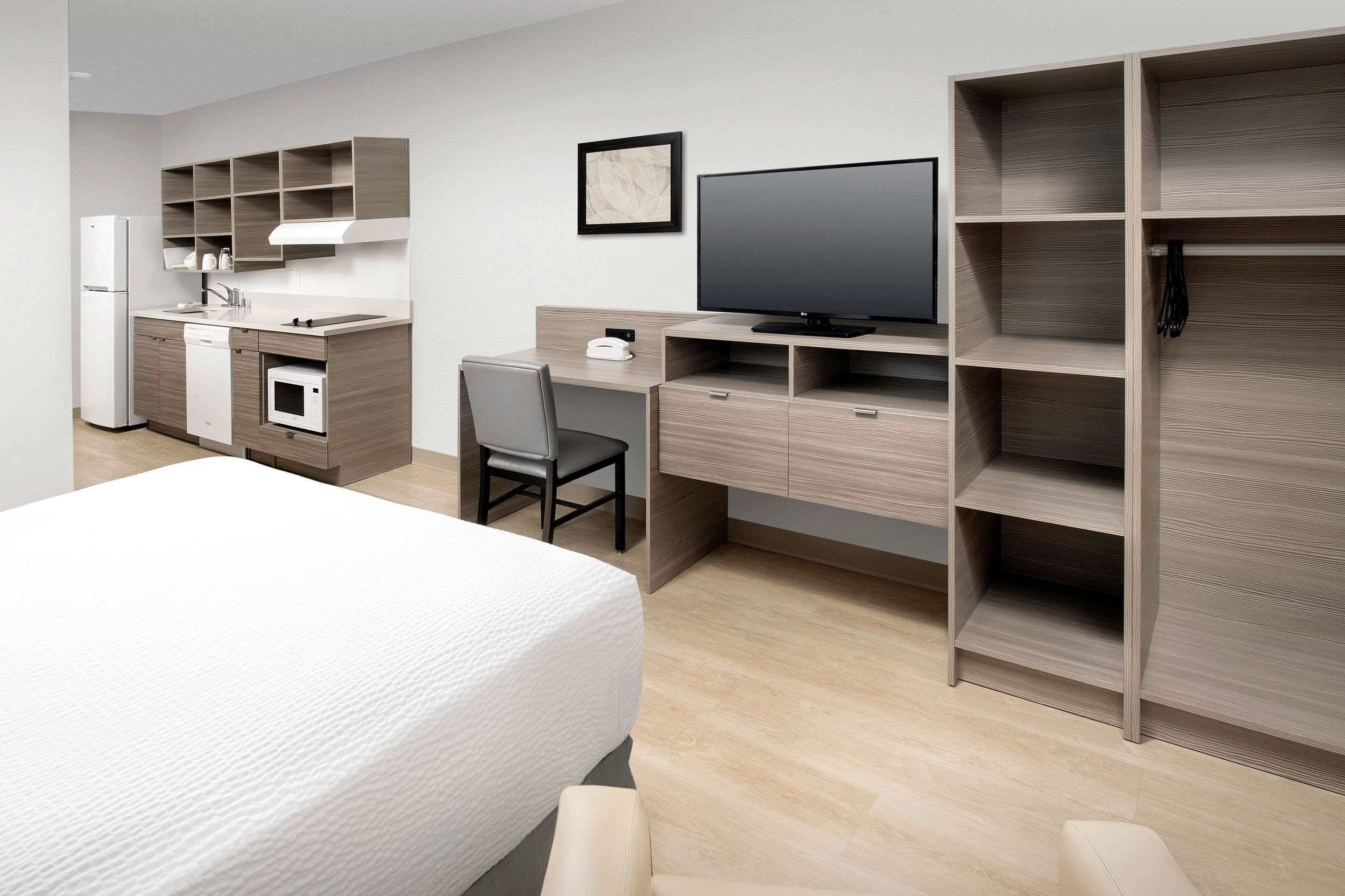 Una habitación de hotel moderna con una cama, un escritorio con computadora, televisor, refrigerador pequeño y armario empotrado.