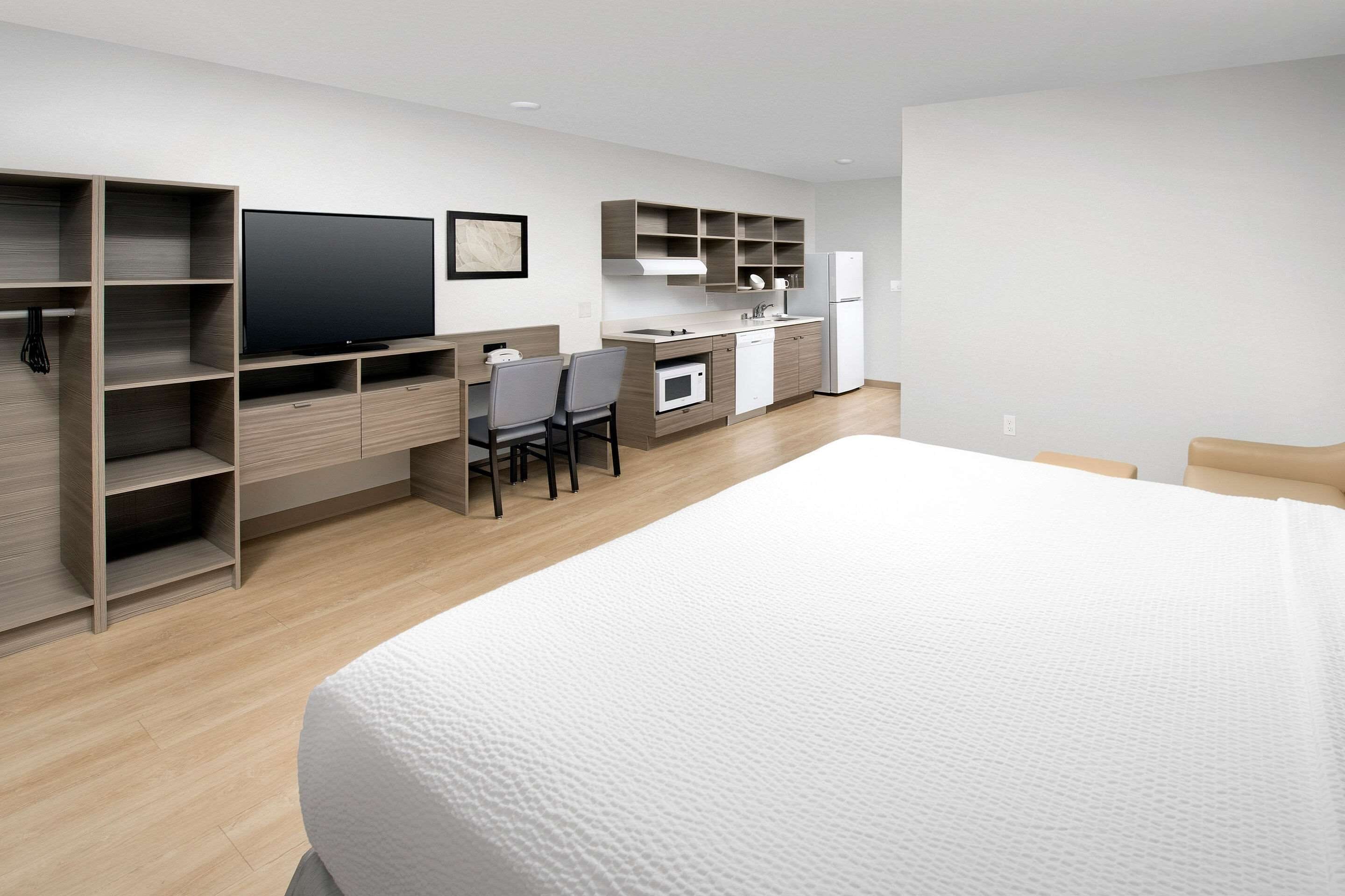 A modern hotel room with a large bed, a desk with chairs, a flat-screen TV, a kitchenette, and built-in shelving.
