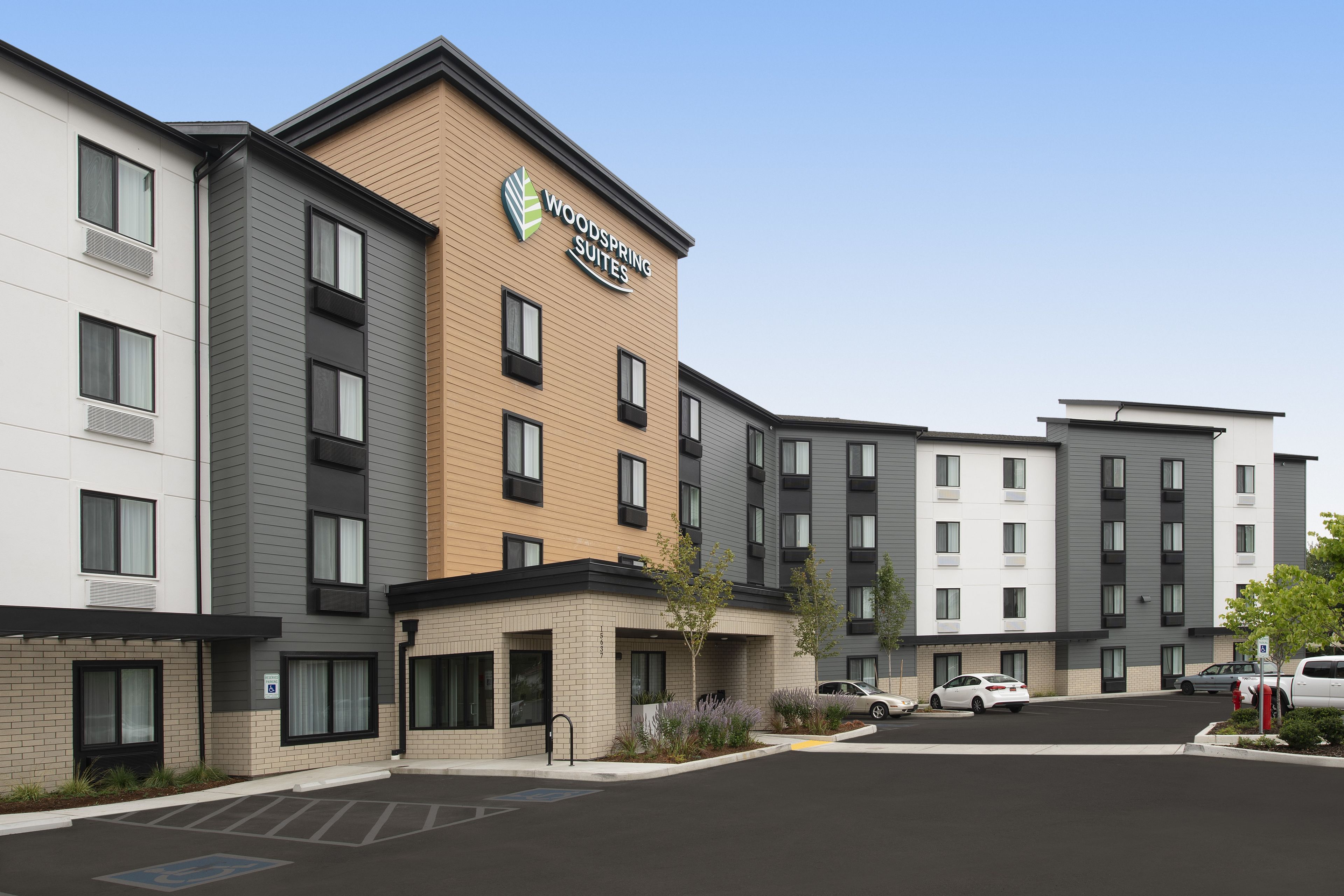 A multi-story hotel with a prominent "Woodspring Suites" sign, a parking lot, and several cars parked in front.