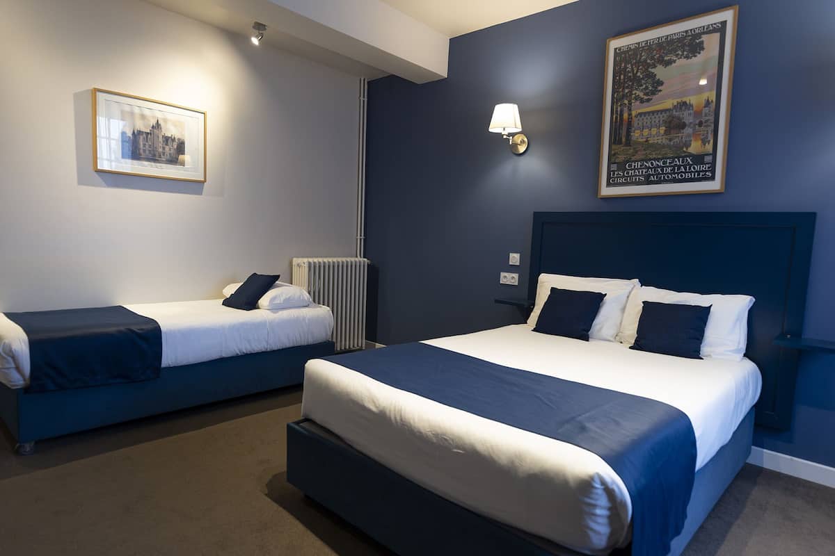 Habitación de hotel con dos camas individuales, una cama grande, un radiador, una lámpara de pared y un cuadro enmarcado.
