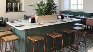 Een eetruimte met hoog plafond, houten balken, een groot raam, een schilderij en een centraal gelegen eiland met krukken.
