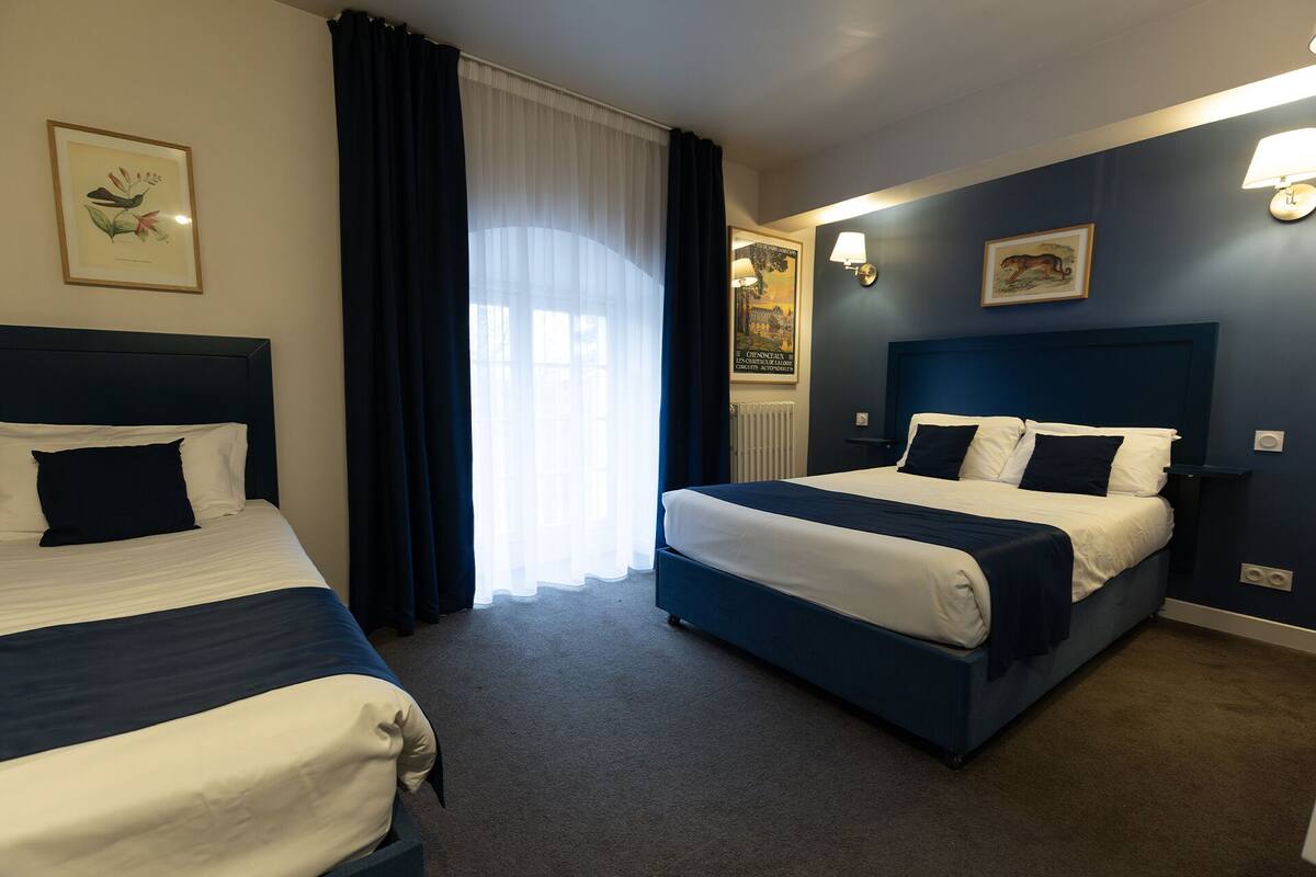 Habitación de hotel con dos camas, un ventanal con cortinas translúcidas e iluminación en la pared.