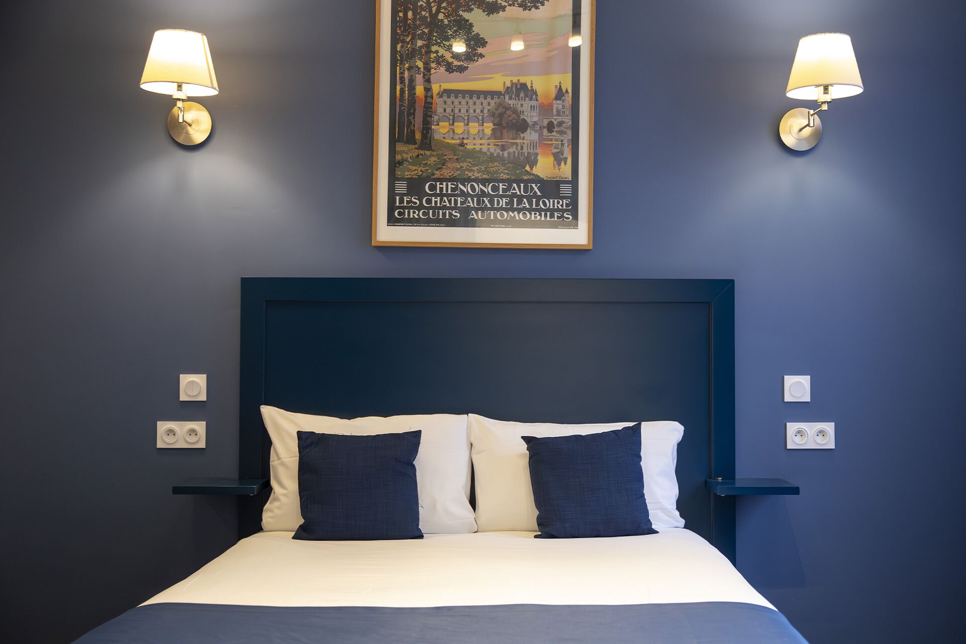 A hotel room with a dark blue headboard, a bed with white and dark blue bedding, two wall-mounted lamps, and a framed picture of a castle on the wall.