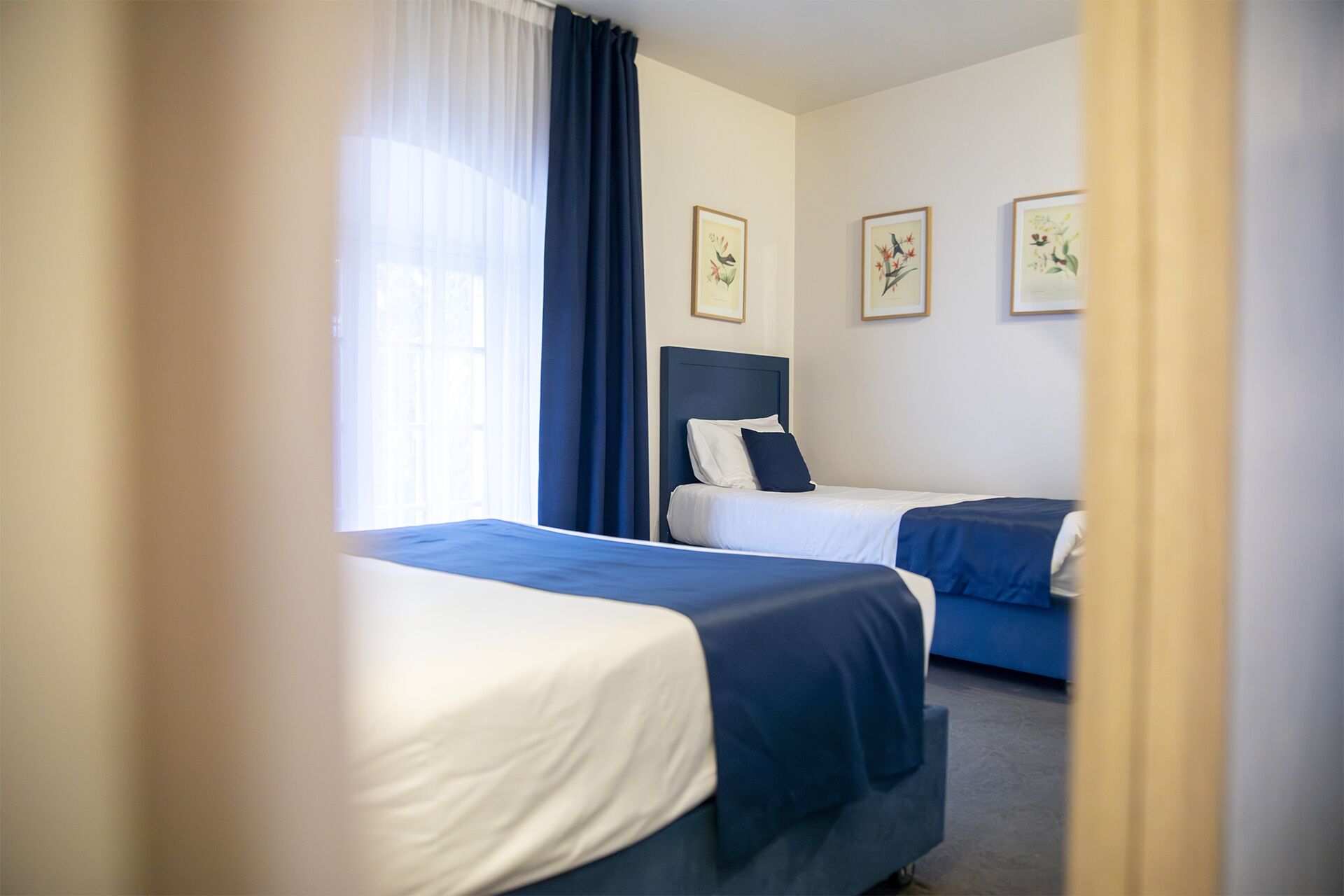 A hotel room with two beds, a large window with sheer curtains, and three framed floral artworks on the wall.