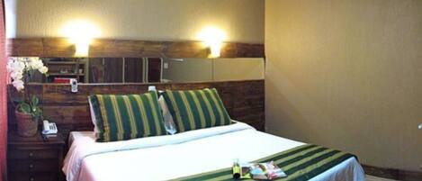 A hotel room with a bed, a nightstand, a lamp, a potted plant, and a wooden headboard with built-in lighting.