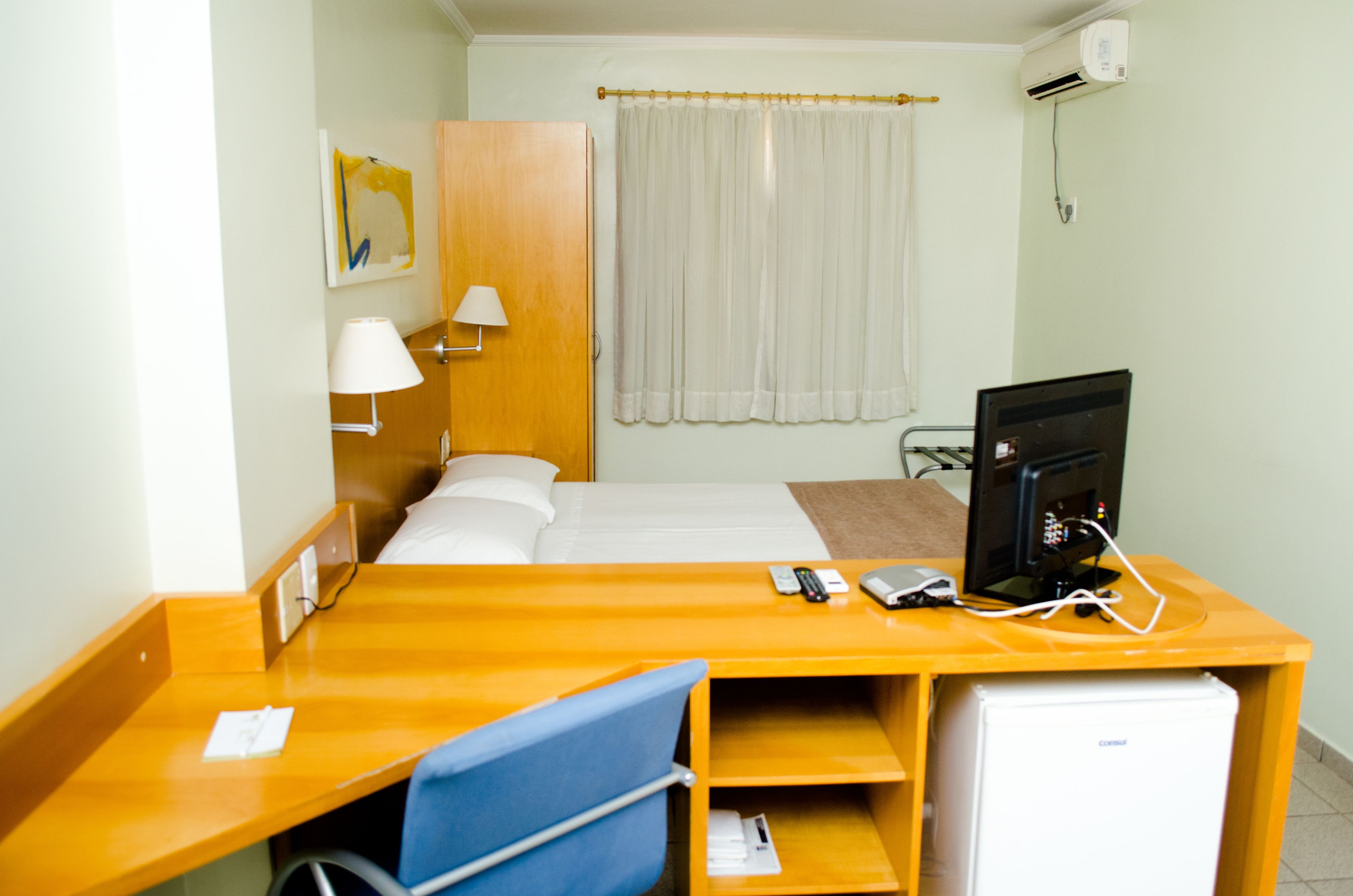 Habitación de hotel con cama, escritorio con computadora, silla, lámpara de escritorio, refrigerador pequeño y ventana con cortinas.