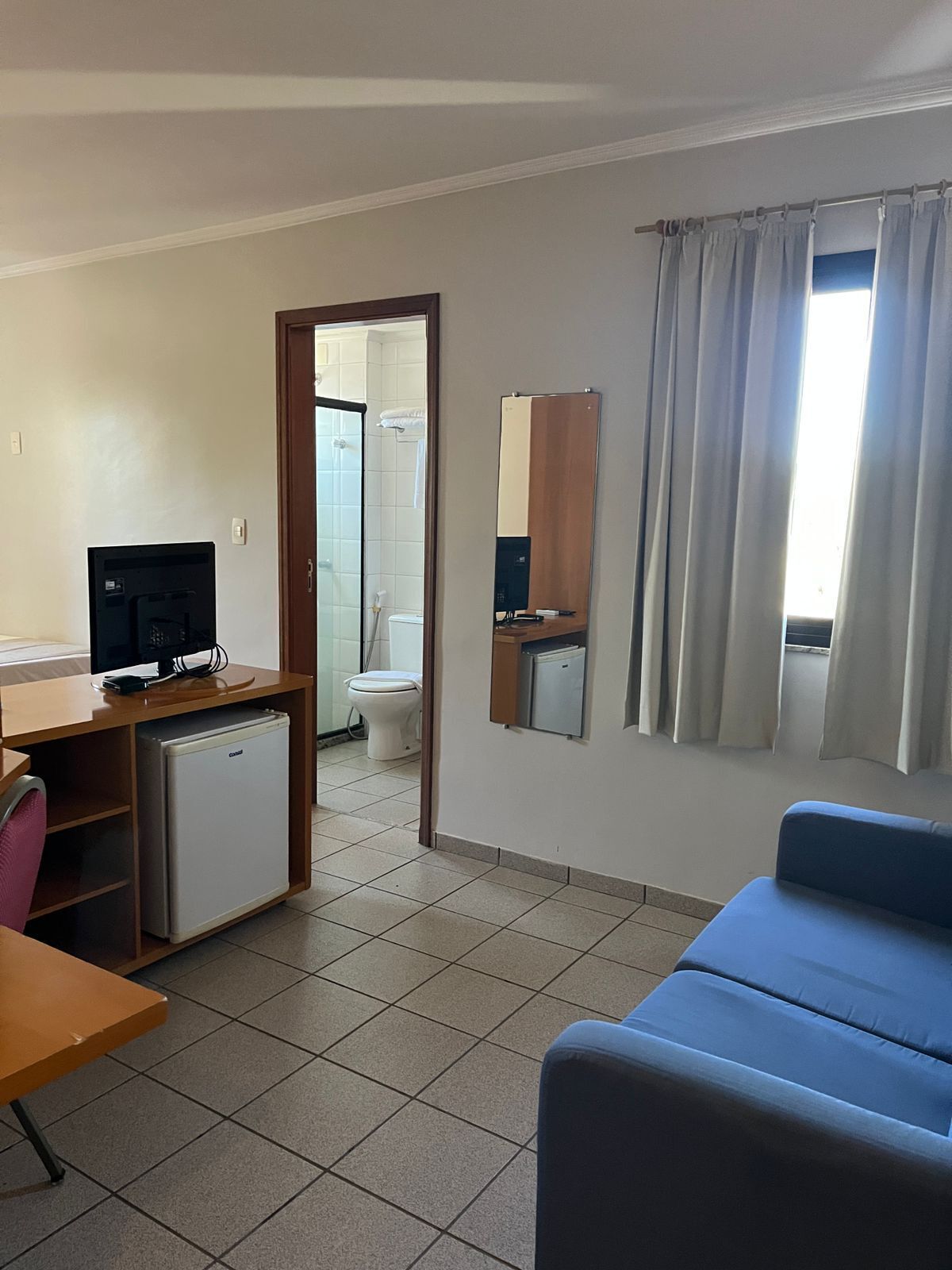 Una habitación compacta con escritorio, refrigerador pequeño, sofá azul, espejo y ventana con cortinas.