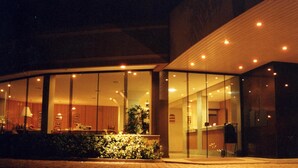 A modern hotel exterior at night with large glass windows, a reception area with a plant, and a brick-paved entrance.