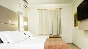 A hotel room with a bed, a bedside table with a lamp, a window with curtains, and a flat-screen TV mounted on the wall.