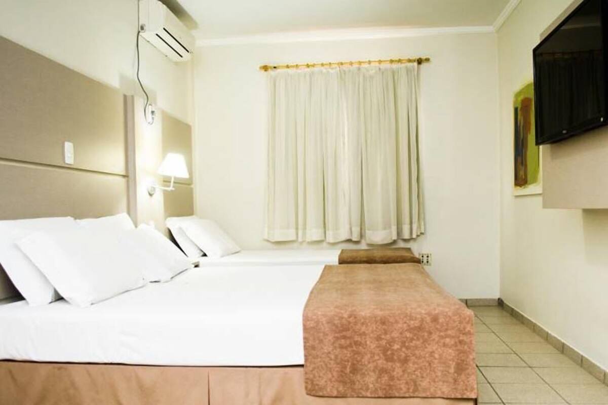 Een hotelkamer met een bed, nachtkastje met lamp, raam met gordijnen en een wandtelevisie.
