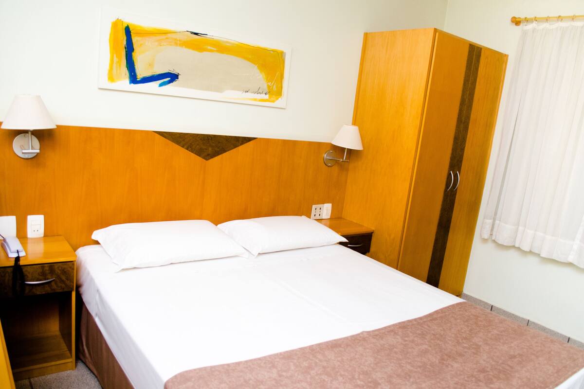 Een hotelkamer met een bed, nachtkastje, lamp, kledingkast en een schilderij aan de muur.