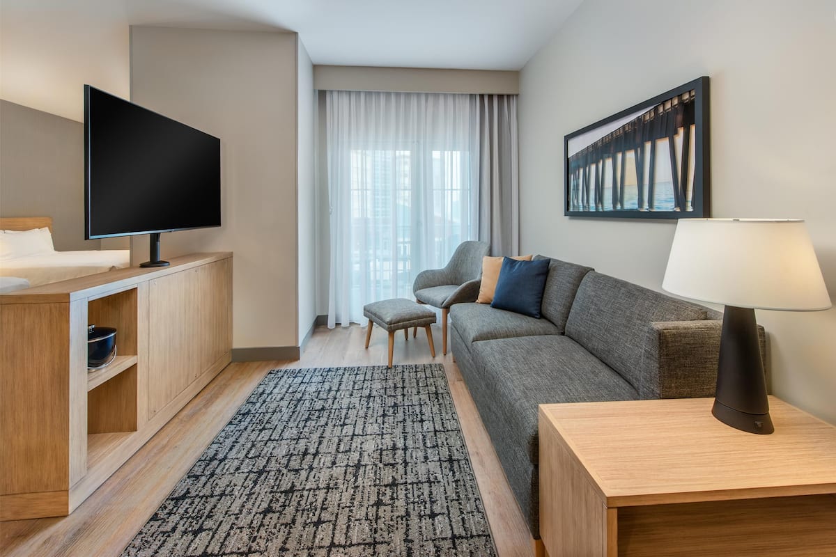 A modern hotel room with a flat-screen TV, a sofa, a small table with a lamp, a wooden cabinet, and a large window with curtains.