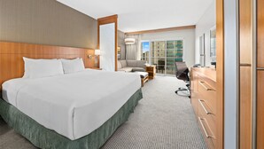 Een hotelkamer met een groot bed, een bureau, een stoel en uitzicht op de stad door grote ramen.