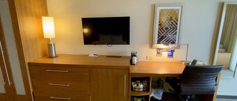 Habitación de hotel con un escritorio de madera, televisión de pantalla plana, cafetera, una silla y una obra de arte enmarcada en la pared.