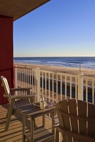 A wooden deck with two Adirondack chairs and a table, overlooking a beach with a railing.