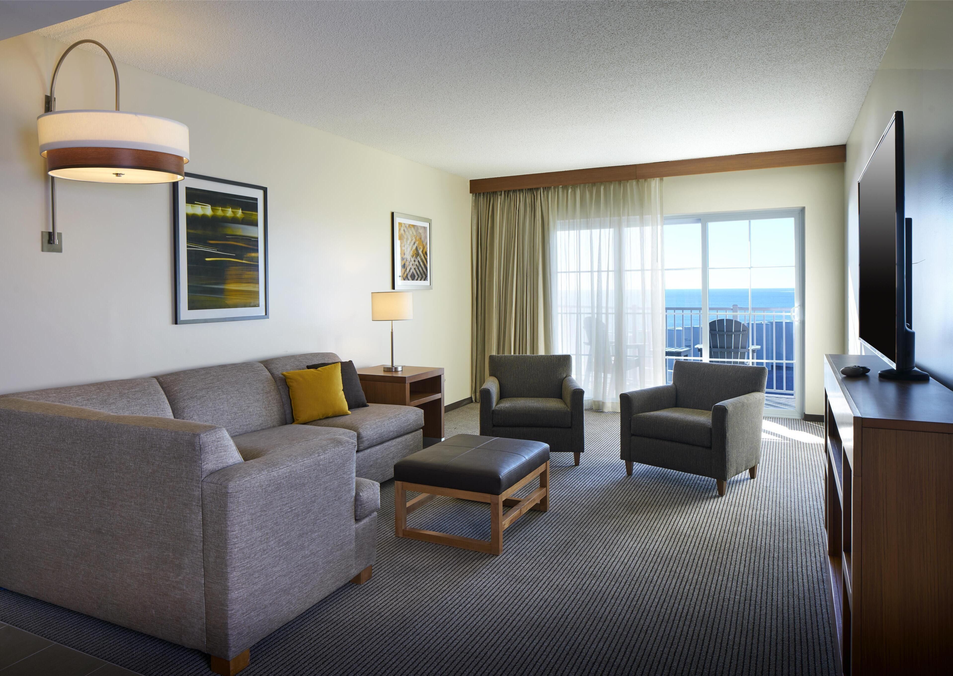Habitación de hotel con sofá, dos sillones, una mesita de centro, televisor, lámpara y un ventanal con vista al océano.