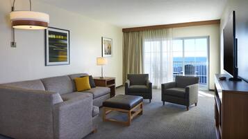 Une chambre d’hôtel avec un canapé, deux fauteuils, une table basse, une télévision, une lampe et une grande fenêtre donnant sur l’océan.