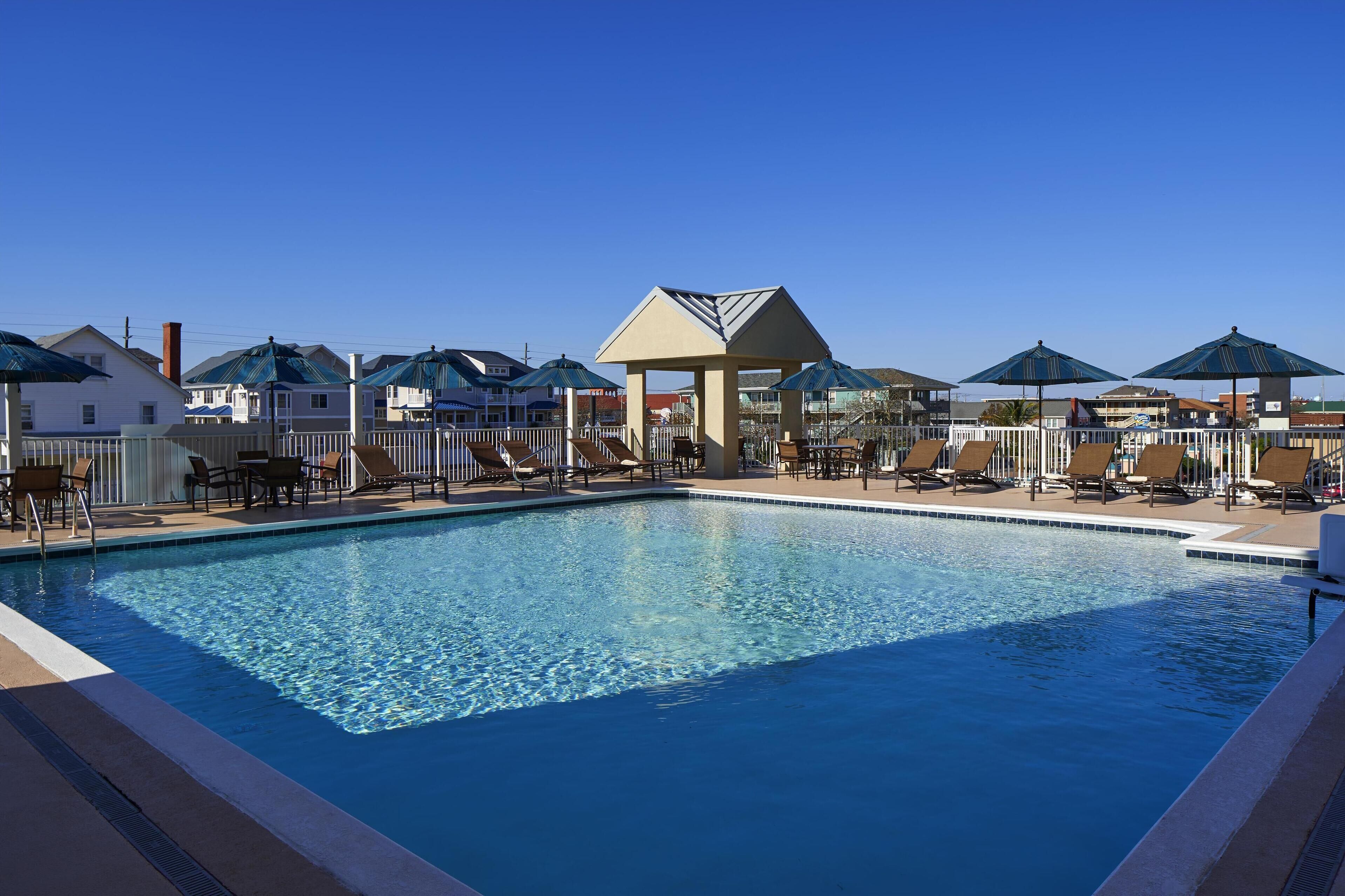 A swimming pool with lounge chairs and umbrellas, surrounded by a fence and buildings.