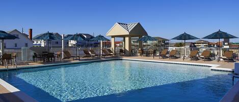 A swimming pool with lounge chairs and umbrellas, surrounded by a fence and buildings.
