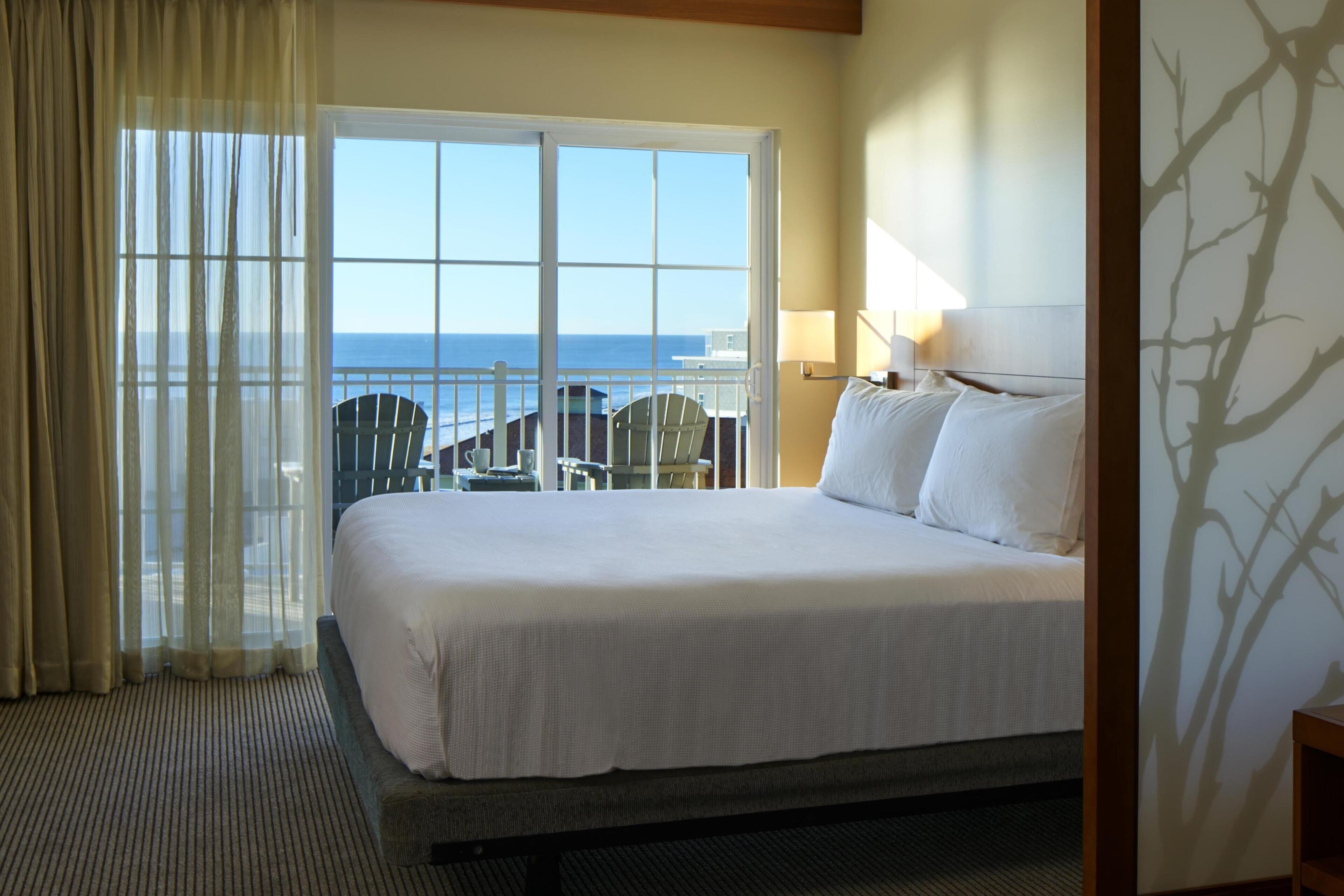 Habitación de hotel con una cama grande, vista al océano y un balcón con sillas.