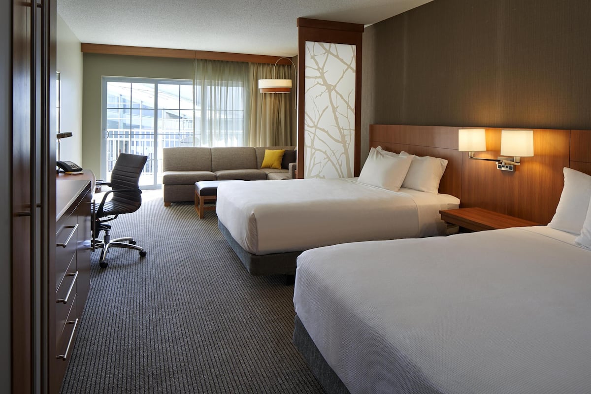 A hotel room with two beds, a desk with a chair, a sofa, and a large window with curtains.