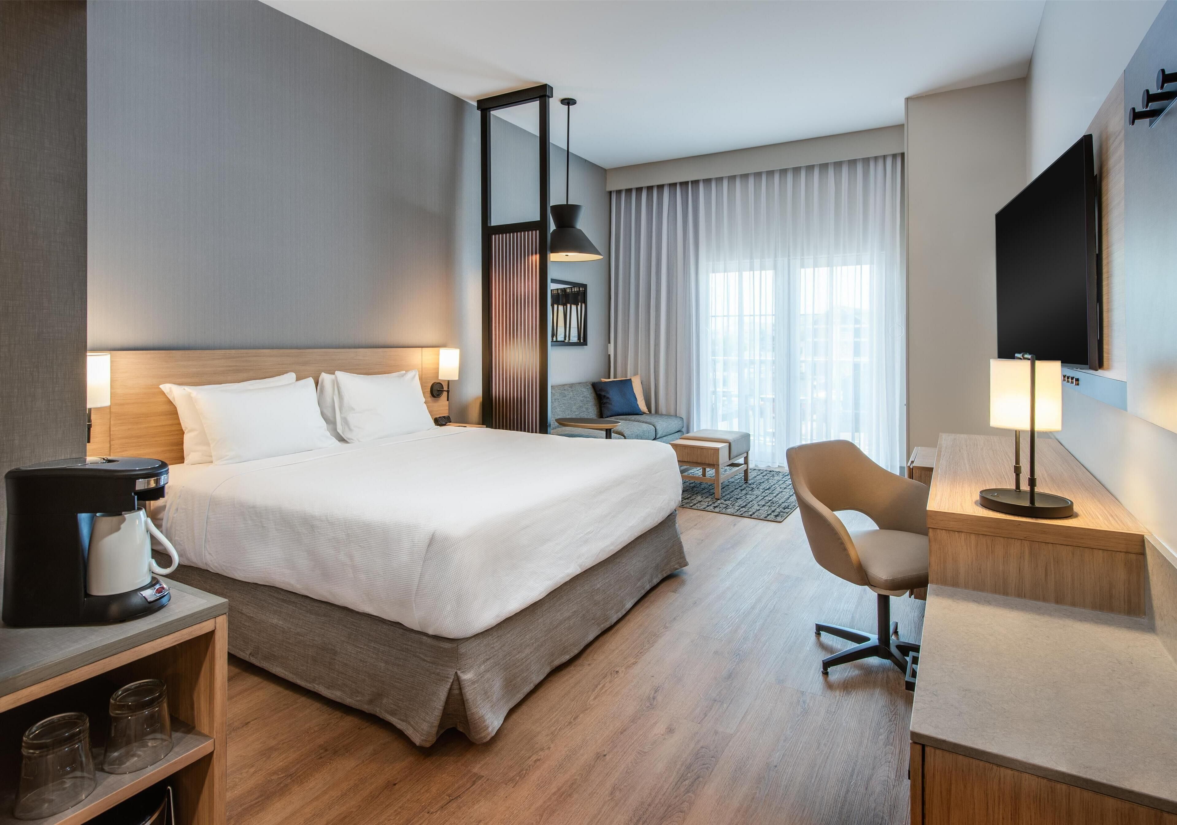 A hotel room with a large bed, a desk with a lamp, a chair, a coffee maker, and a TV.