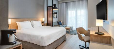 A hotel room with a large bed, a desk with a lamp, a chair, a coffee maker, and a TV.