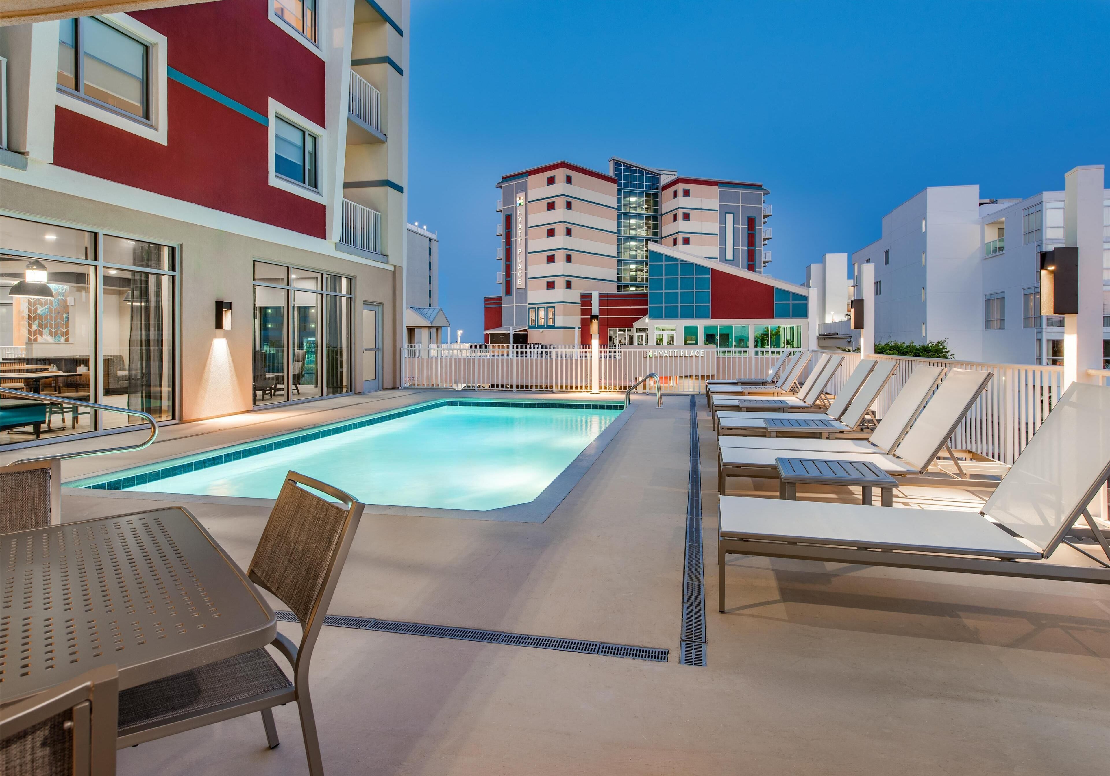 Ein Poolbereich mit Liegestühlen und einem Tisch, ein Gebäude mit roter und weißer Fassade und ein klarer Himmel.