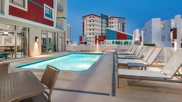 Ein Poolbereich mit Liegestühlen und einem Tisch, ein Gebäude mit roter und weißer Fassade und ein klarer Himmel.