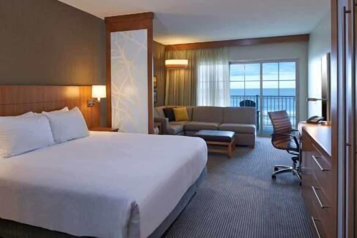 A hotel room with a large bed, a sofa, a desk, and a chair. There is a window with a view of the ocean.