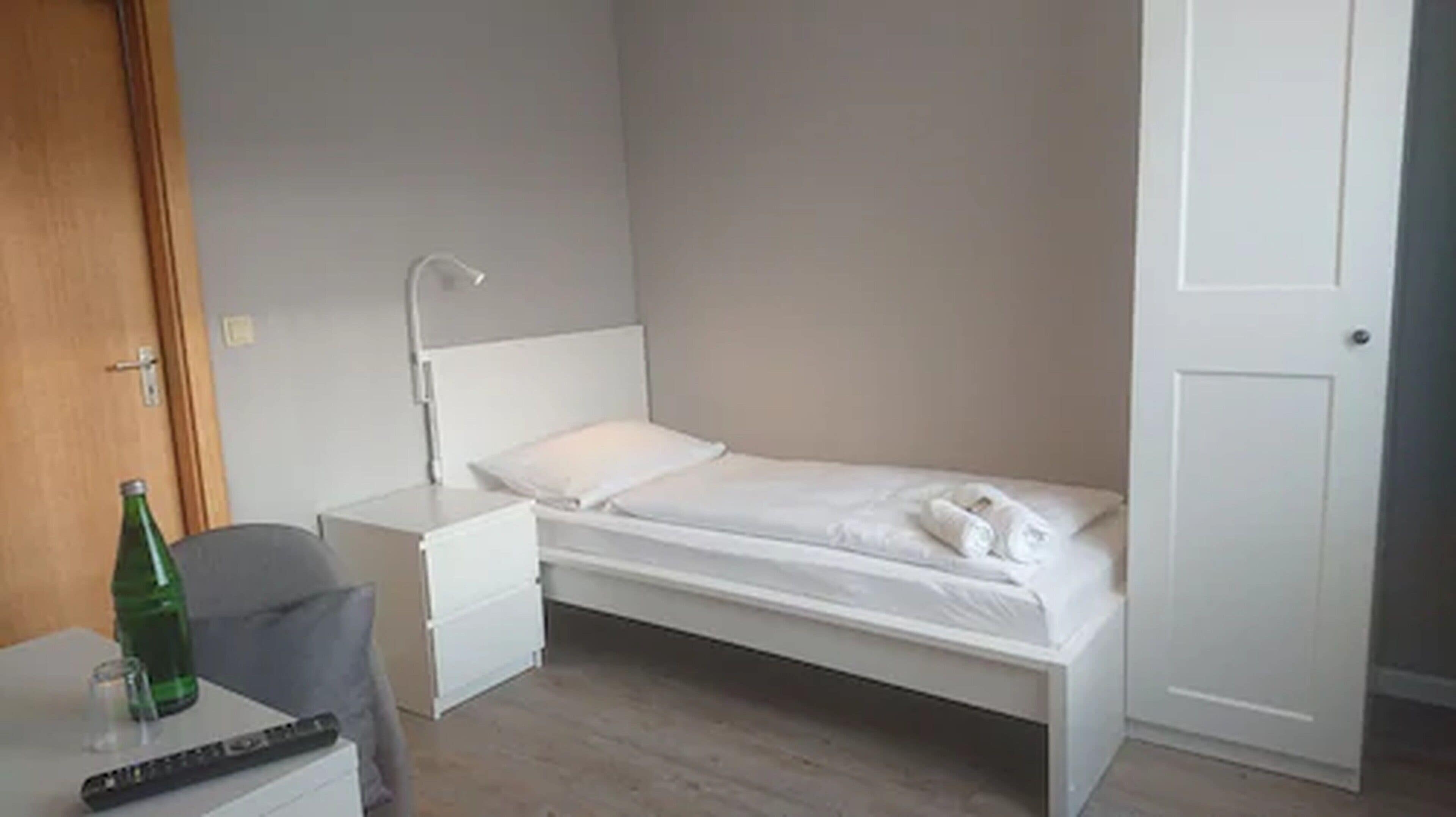 A small, single-bed room with a wooden floor, a white bed, a bedside table with a lamp, and a chair.