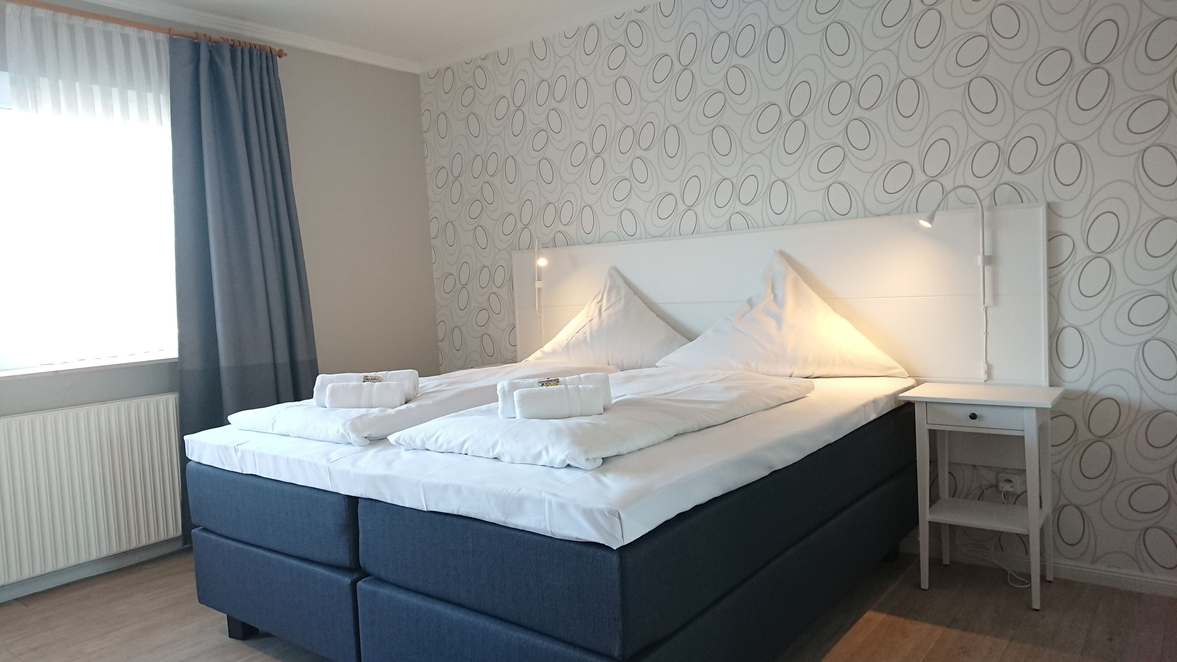 Un lit bien fait, avec du linge de lit blanc et deux serviettes, une table de chevet avec une lampe et un mur décoré d’un motif.